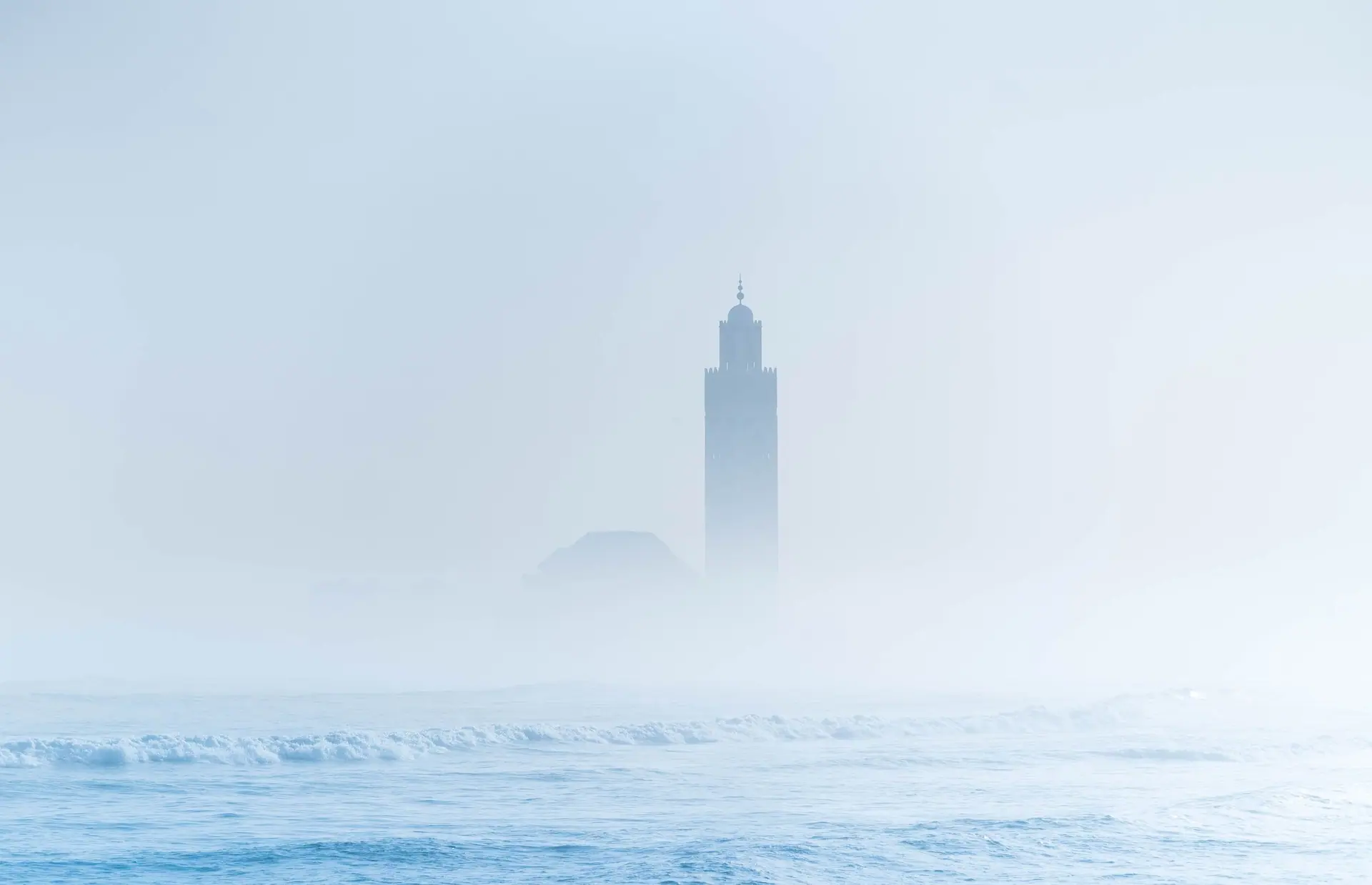 View of Hassan II Mosque in Casablanca shrouded in mist, with ocean waves in the foreground.