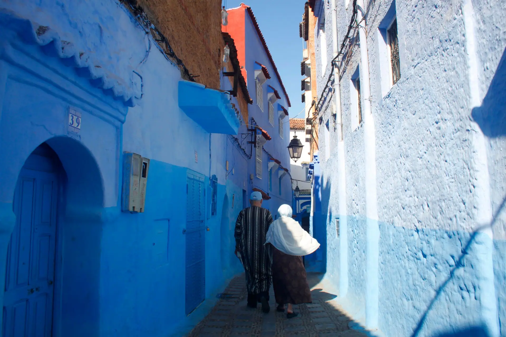 A couple walks through the vibrant blue-painted alleyways of Chefchaouen, Morocco, under the midday sun.