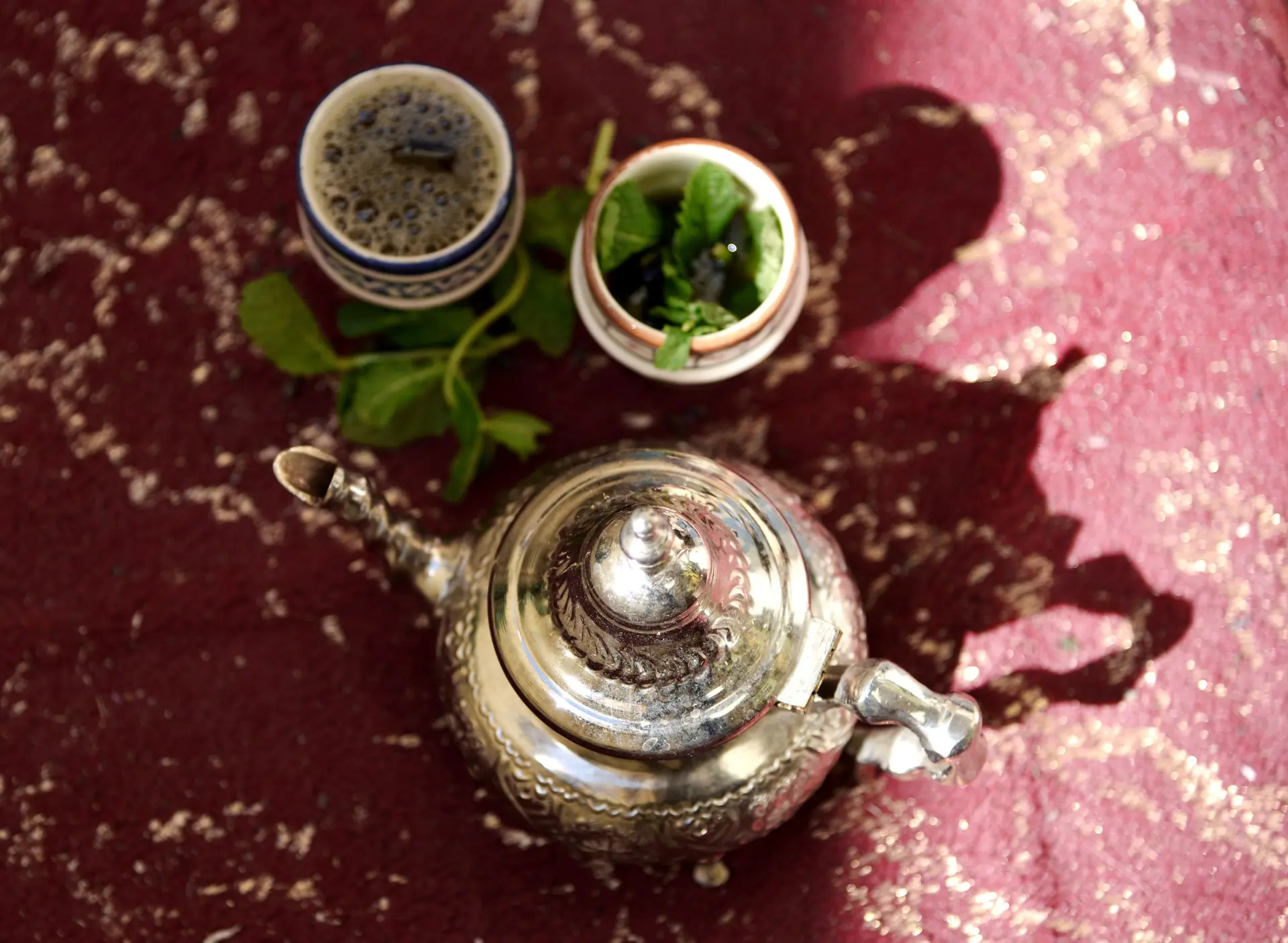 A silver teapot and teacups with mint on a textured Moroccan carpet in sunlight.